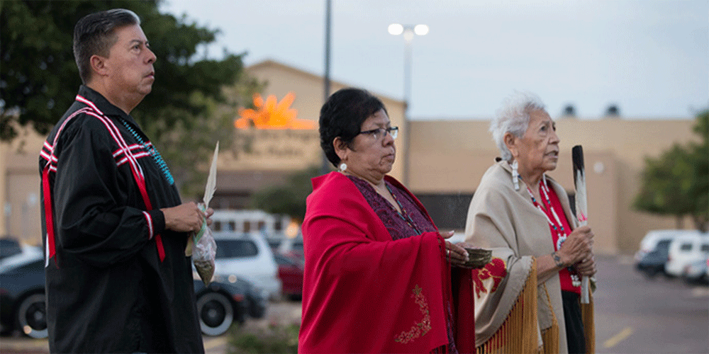 Bishop David Wilson (left) and the Rev. Donna Pewo (center) of The United Methodist Church’s Oklahoma Indian Missionary Conference join with Native American scholar Henrietta Mann in a prayer service for immigrant children held at the Casa Padre detention center, visible behind them, in Brownsville, Texas, in 2018. File photo by Mike DuBose, UM News.